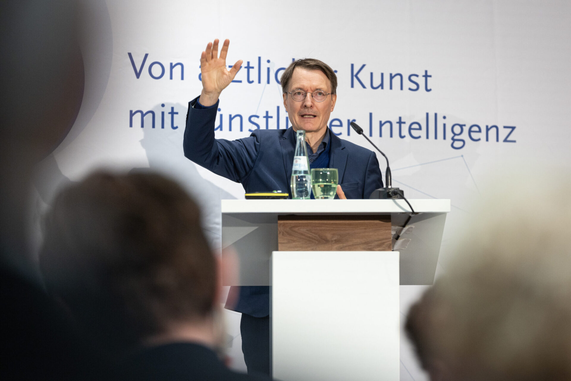 Karl Lauterbach spricht bei der Veranstaltung ‚BAEK im Dialog‘ der Bundesärztekammer. Er steht hinter einem Rednerpult, hebt die Hand und richtet sich an das Publikum. Im Vordergrund sind unscharfe Zuhörer zu sehen. Ein typisches Beispiel für Event- und Kongressfotografie, das einen Vortrag während eines medizinischen Fachdialogs festhält.